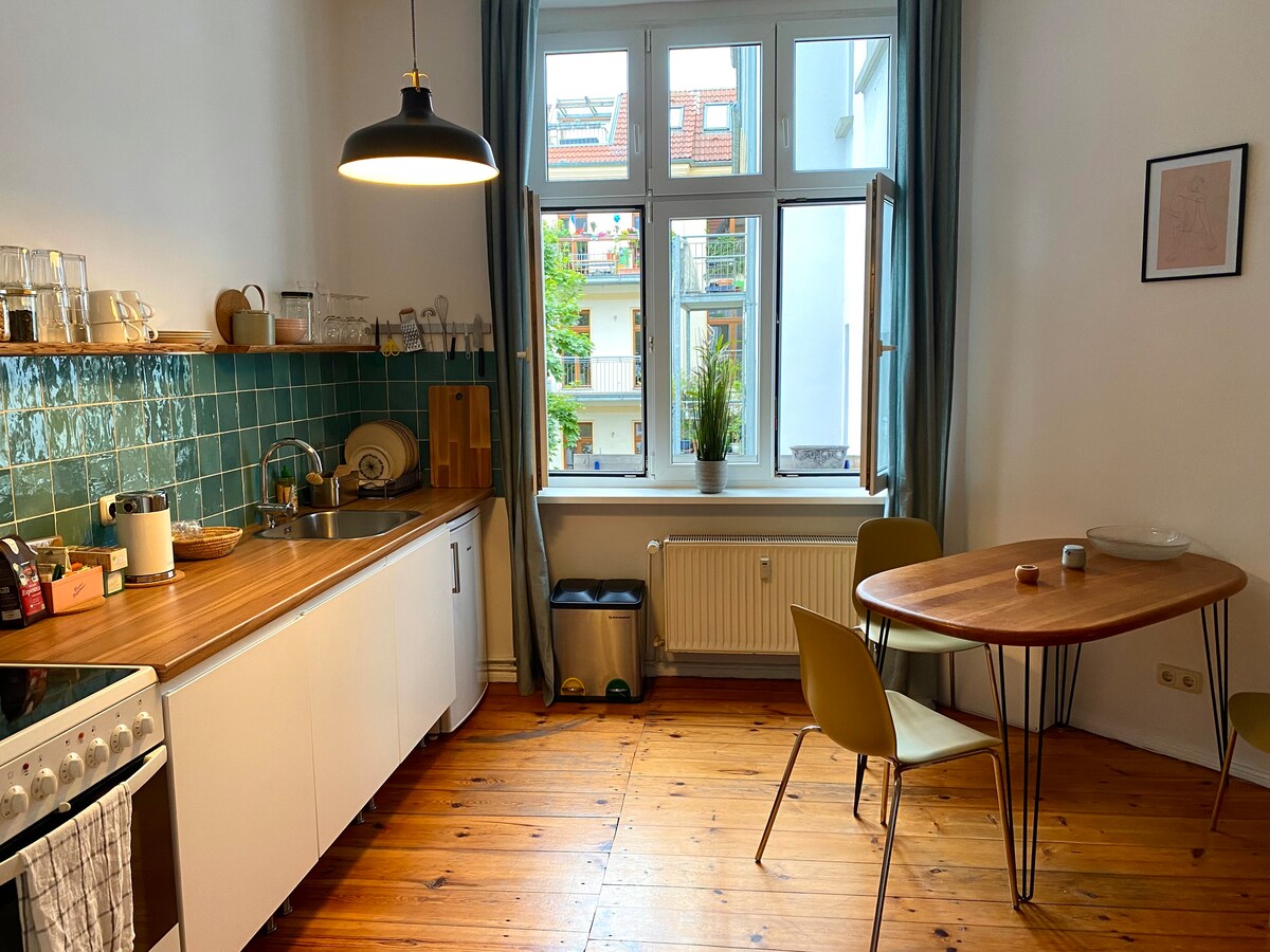 A well-equipped kitchen area is highlighted by a wooden countertop and teal tiled backsplash. Sunlight enters through large windows, illuminating the space. A round dining table with two chairs stands ready for use, and a kitchen sink and stove are visible, creating a functional environment.