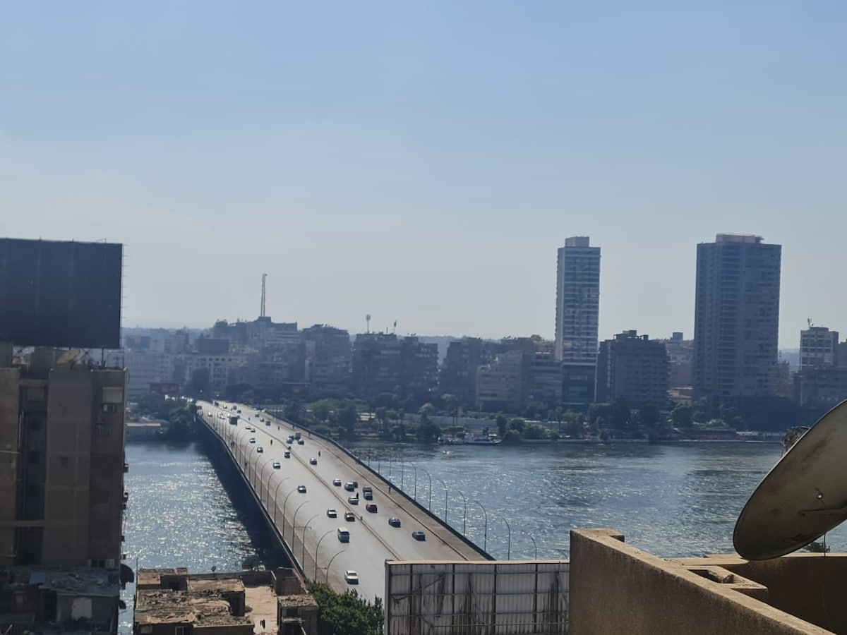 A panoramic view of the Nile River is captured from an elevated position. The scene features a bridge with vehicles traveling across it, flanked by buildings along the riverbank under a clear sky.