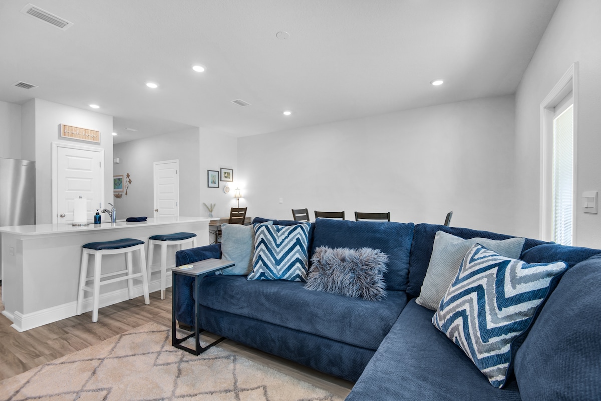 A modern living area features a large blue sectional sofa adorned with decorative pillows. A textured rug lies on the floor, complementing the light wood tones. In the background, a kitchen island with bar seating is visible, along with dining tables and framed artwork.