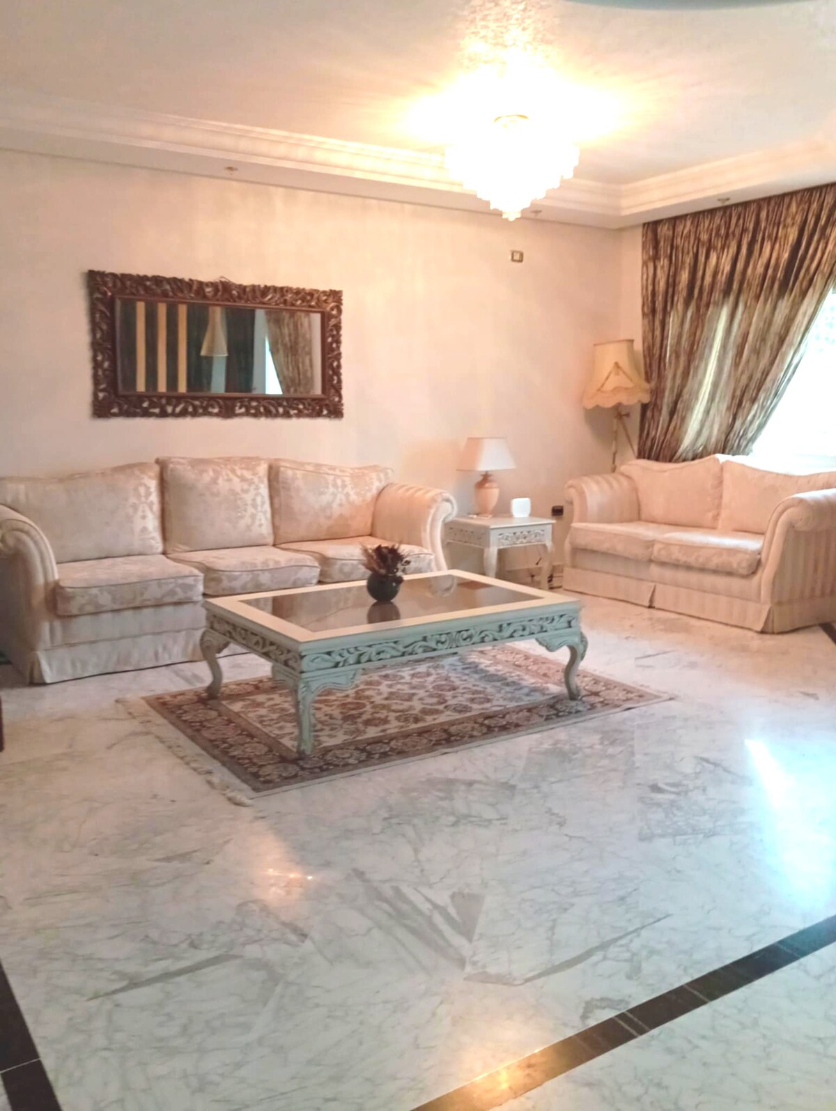 A bright living area features elegant seating with cream-colored sofas and a glass coffee table. A decorative mirror adorns the wall above the table, reflecting soft light. Natural light enters through a window draped with patterned curtains, complementing the marble floor beneath.