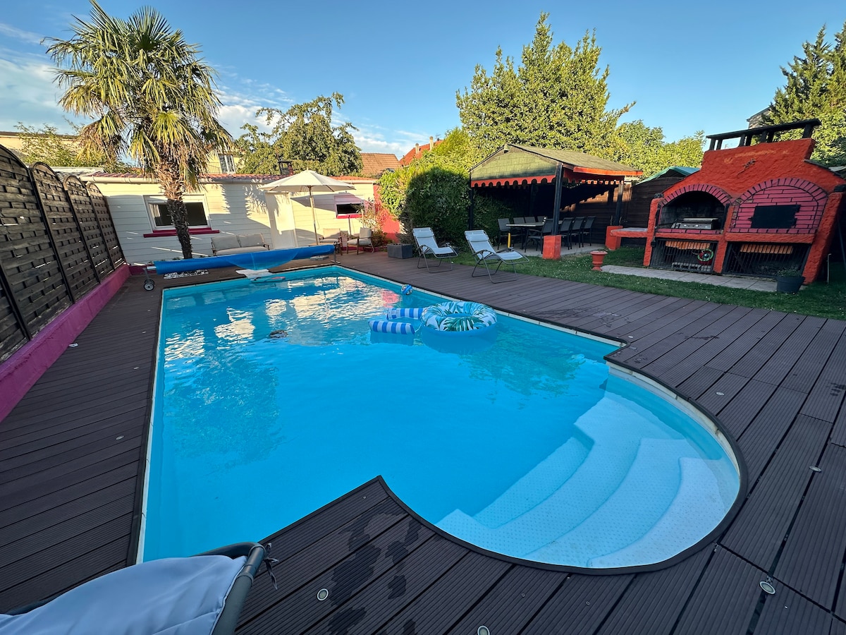 The outdoor space features a shaped pool surrounded by wooden decking. Lounge chairs are positioned near the pool, and a shaded area with outdoor seating is visible. Lush greenery and a barbecue pit enhance the appeal of this tranquil garden setting.