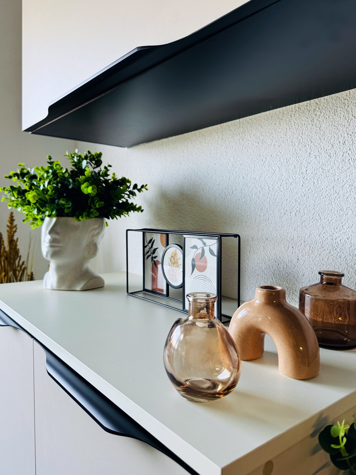 A modern shelving unit is presented, featuring a collection of decorative items. A small potted plant adds a touch of greenery, while several glass vases in varying shapes and colors are displayed alongside a framed arrangement of art. The clean white surface emphasizes the decor.