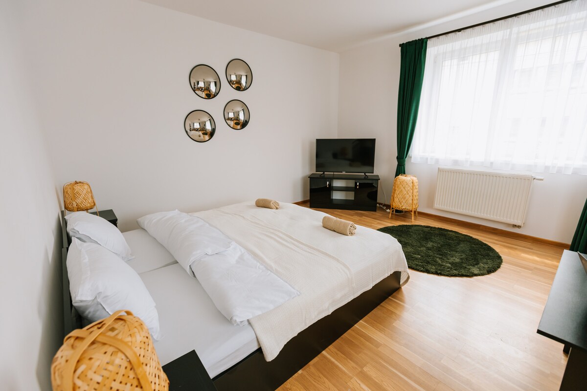 A spacious bedroom features a large, configured bed with soft linens. Three round mirrors adorn the white wall, reflecting light. A TV is positioned on a dark cabinet, while two woven lamps provide gentle illumination by the window, draped with sheer curtains.