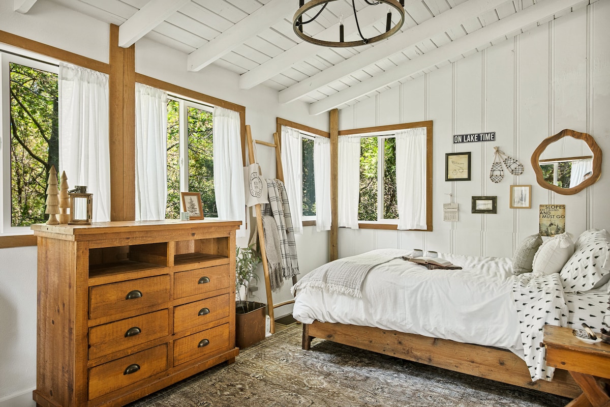 The primary bedroom is illuminated by sunlight streaming through multiple windows draped with white curtains. A spacious bed is adorned with simple bedding, while a wooden dresser adds rustic charm. Subtle decor elements enhance the room's inviting ambiance, complemented by views of the surrounding lush forest.