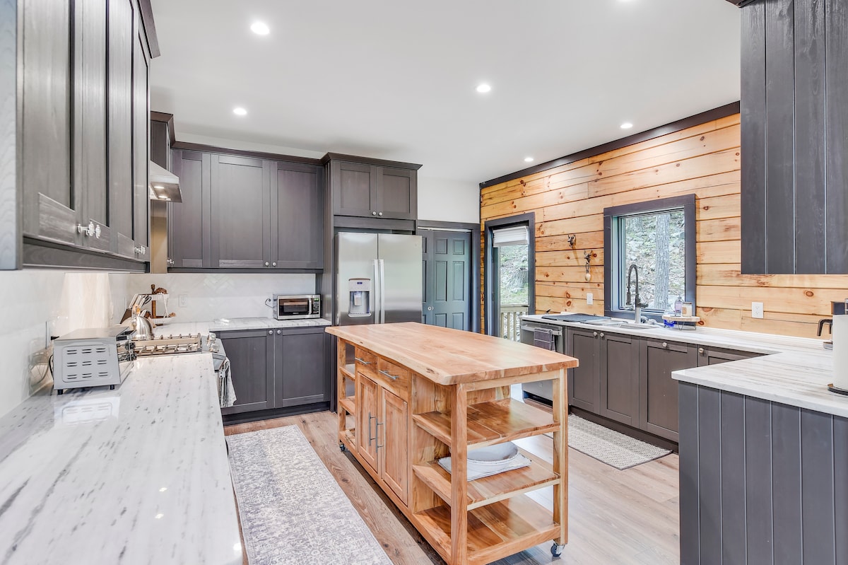 A modern kitchen is presented with dark cabinetry and marble countertops. An island with a light wood finish creates a central workspace. Large windows provide natural light, while stainless steel appliances and a distinct wooden accent wall contribute to the space's inviting ambiance.