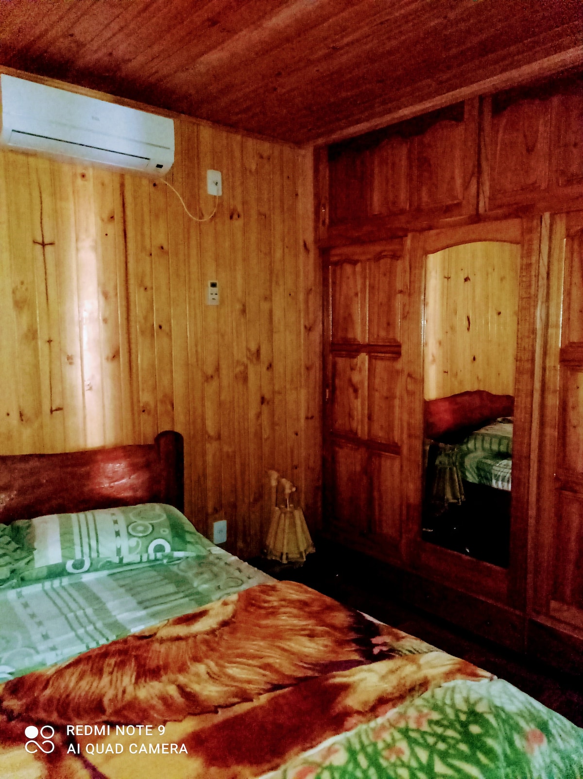 A cozy bedroom features warm wooden paneling and a spacious bed adorned with a vibrant green and patterned blanket. Natural light enhances the space through a large mirror, while an air conditioning unit and a small decorative lamp are visible.