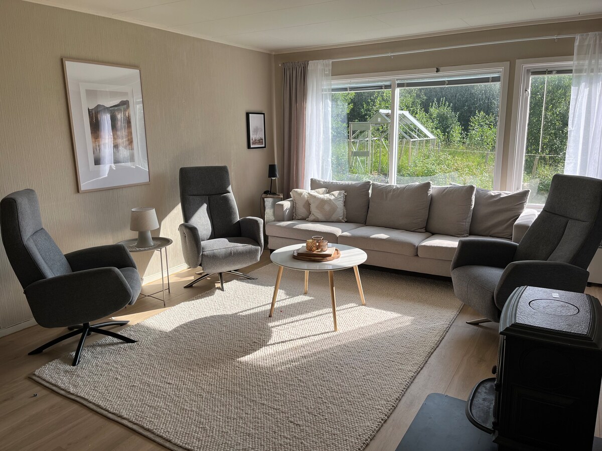 A bright living room is featured, with a cozy gray sofa facing the large windows that allow natural light to fill the space. Two modern armchairs are positioned near a round coffee table, enhancing the inviting atmosphere. A textured area rug rests on the wooden floor.