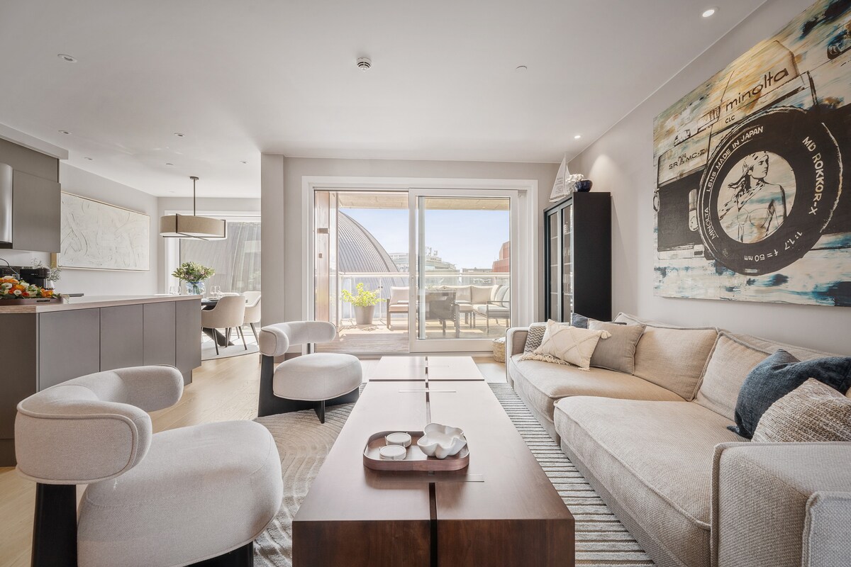 A spacious living area is presented, featuring a light-colored sofa and modern armchairs. A sleek wooden coffee table sits in the center, complemented by decorative items. Floor-to-ceiling windows allow natural light, revealing an outdoor terrace beyond. Neutral tones create a calming atmosphere.