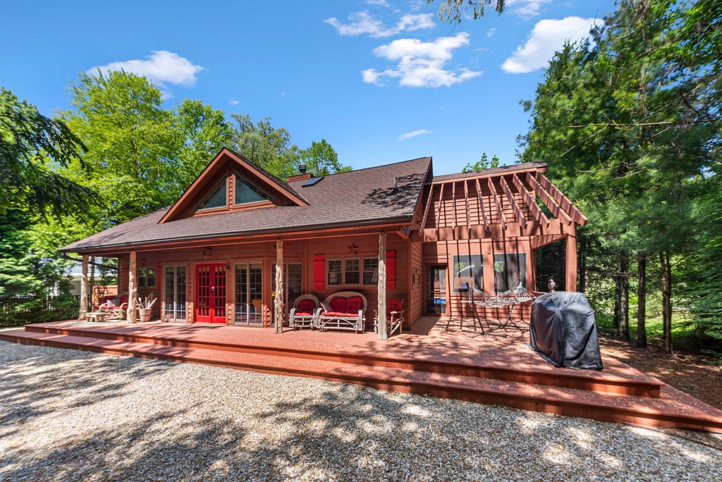 A charming exterior of a red-accented house is showcased, featuring a spacious deck with seating areas and a barbecue grill. Lush greenery surrounds the property, enhancing the sense of privacy and tranquility. Tall trees provide shade, while a clear blue sky completes the inviting scene.