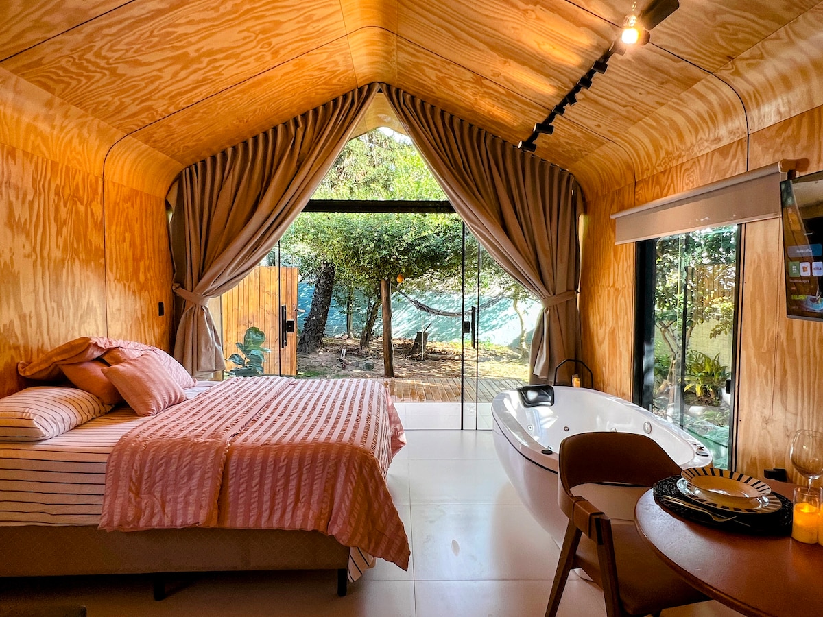 A cozy bedroom features a spacious bed covered with a soft pink blanket. A modern hot tub is positioned nearby. Floor-to-ceiling glass doors open to a private outdoor area with lush greenery, allowing natural light to fill the space.