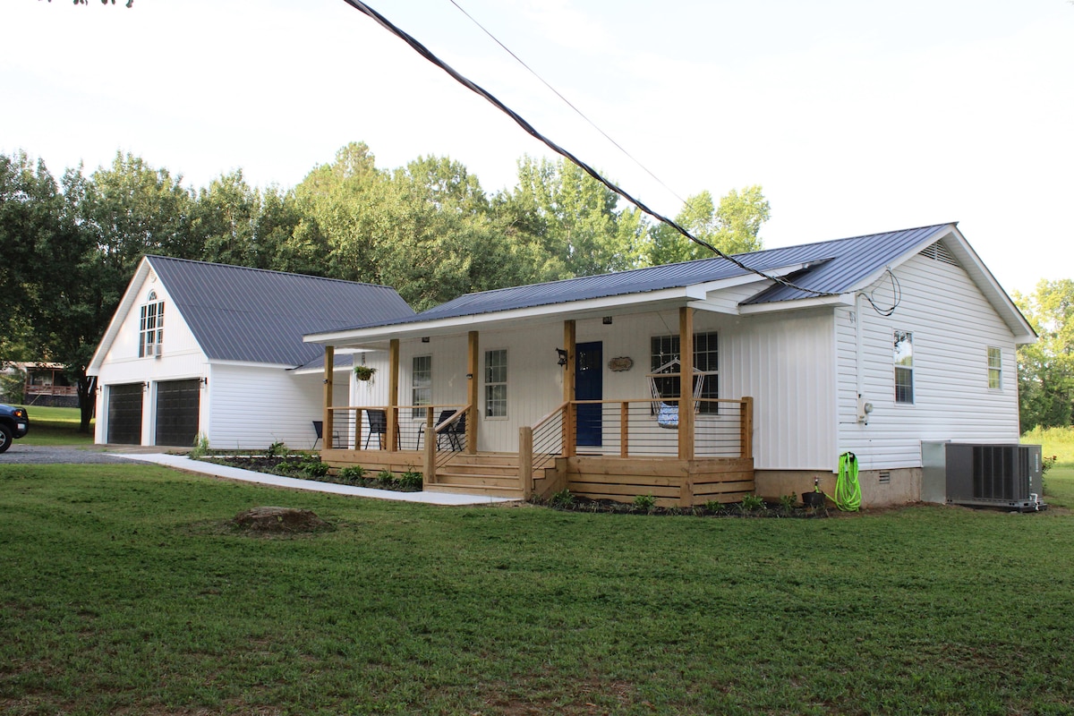 A single-story home is shown with a light-colored exterior and a metal roof. A wooden porch, featuring railings, invites relaxation with seating options. Adjacent, a garage complements the property, surrounded by open grass areas and mature trees in the background.