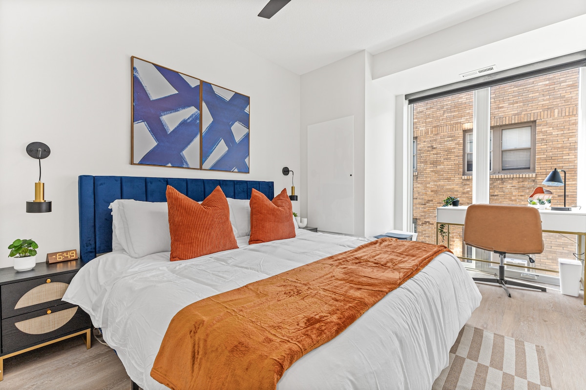 A cozy bedroom features a spacious bed adorned with two orange pillows and a soft throw blanket. Vibrant wall art is displayed above the headboard. Natural light streams through large windows, illuminating a desk and chair positioned nearby.