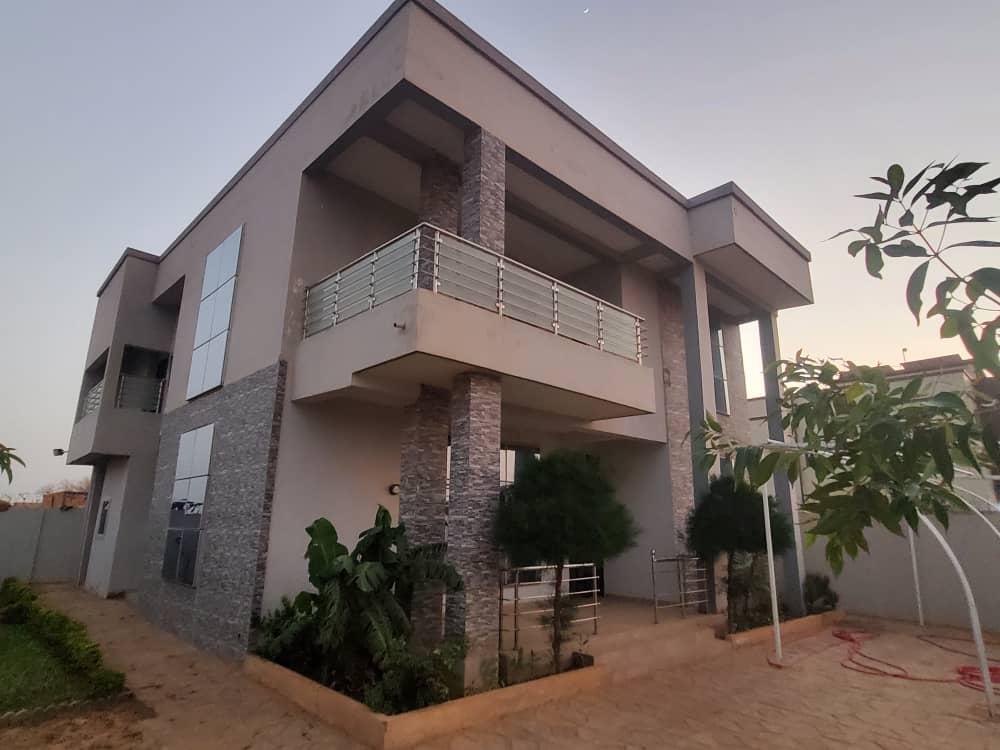 The modern villa features a contemporary architectural design with a mix of stone and smooth surfaces. A spacious balcony provides ample outdoor space, surrounded by plants. The exterior is well-maintained, showcasing a serene garden area and a pathway leading to the entrance.