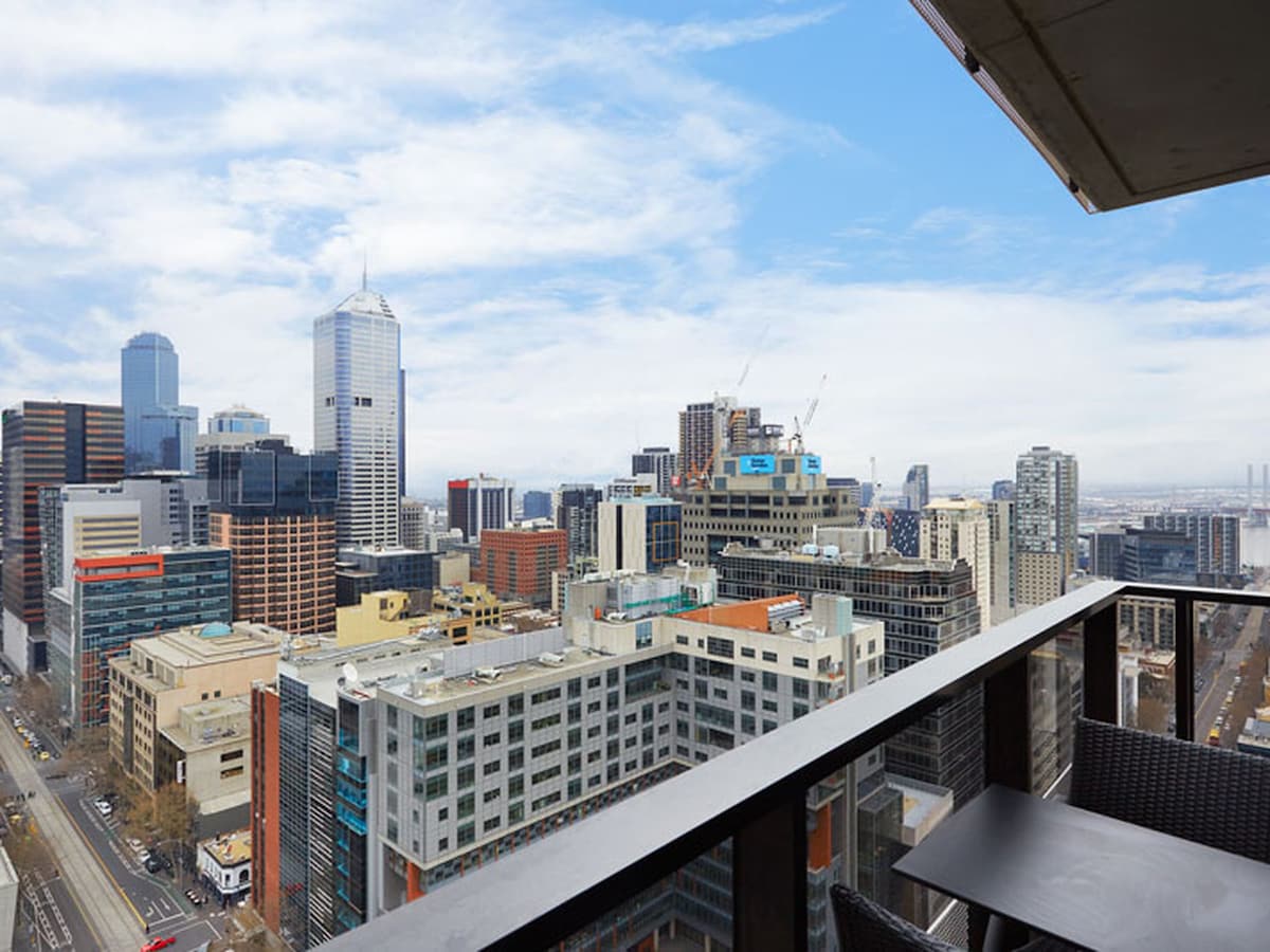 A private balcony overlooks the dynamic Melbourne skyline, showcasing high-rise buildings and bustling city life. The panoramic view captures a blend of modern architecture against a light blue sky, enhancing the urban experience from this 20th-floor studio.