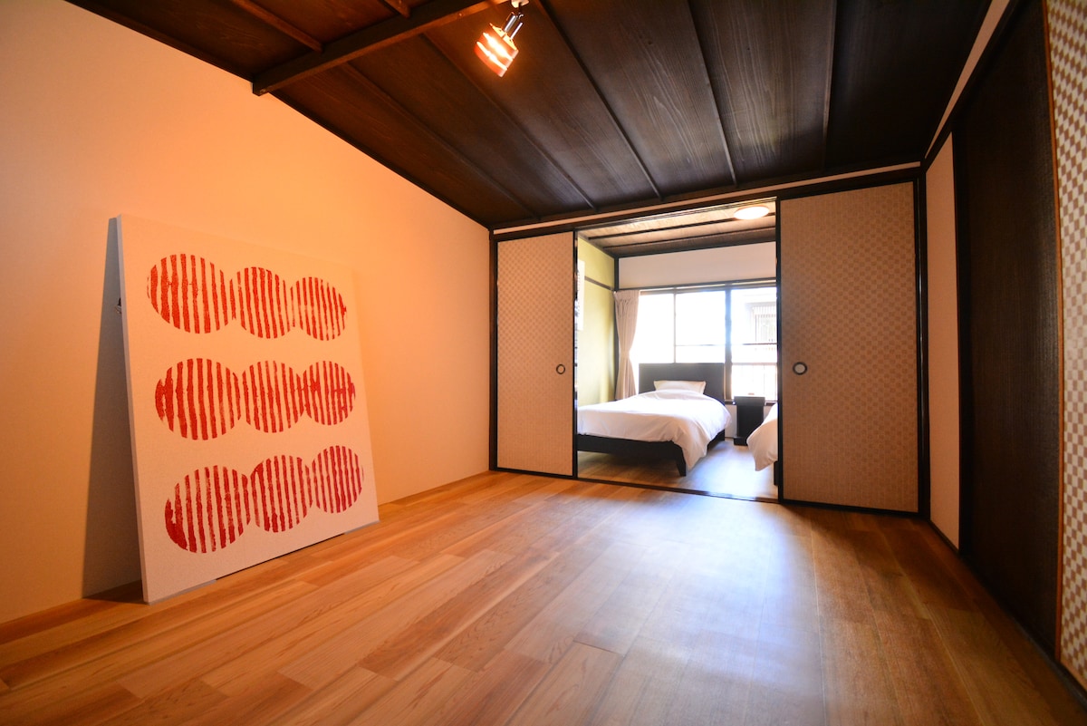 A spacious room displays a striking piece of modern art against a light-colored wall. Natural wood flooring complements the traditional sliding doors that open to a sleeping area with a visible bed. Soft light enters through the window, enhancing the relaxed ambiance.