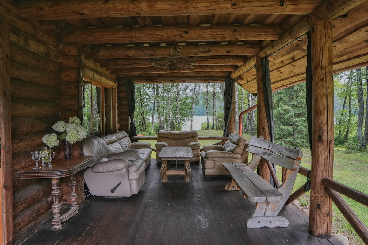 A covered outdoor seating area features a mix of comfortable leather sofas and a wooden table, surrounded by natural wood beams. A view of the lake and trees is visible through large windows, creating a serene atmosphere for relaxation.