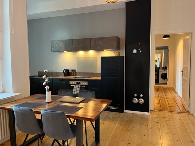 A modern kitchen and dining area is presented, featuring a wooden dining table with gray chairs. The kitchen is equipped with dark cabinetry, a built-in oven, and essential appliances. Warm lighting accents the space, enhancing the neutral color palette of the walls and flooring.
