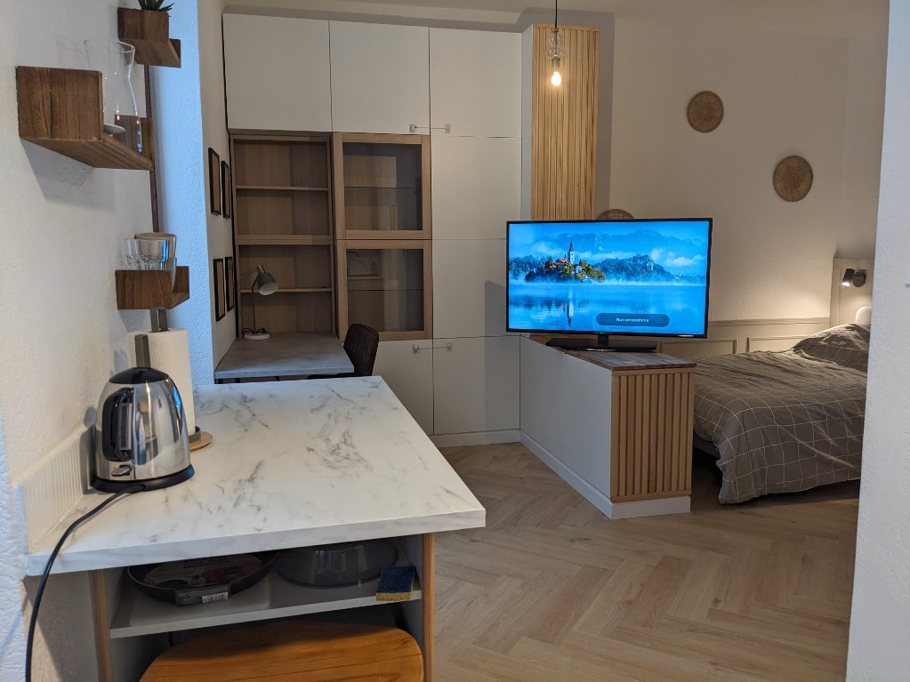 A modern living space is shown, featuring a large flat-screen TV positioned in front of a cozy sleeping area with a neatly made bed. A kitchenette area is visible with a marble countertop and an electric kettle, complemented by wooden shelves and warm lighting.