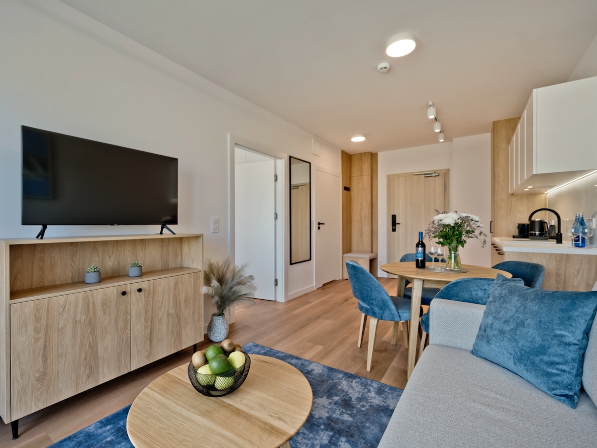 A modern living area is showcased, featuring a cozy sofa and a round dining table surrounded by stylish chairs. A television is mounted on a light wood cabinet, while fresh flowers and a bowl of fruit add a touch of color. Light wood finishes enhance the bright atmosphere.