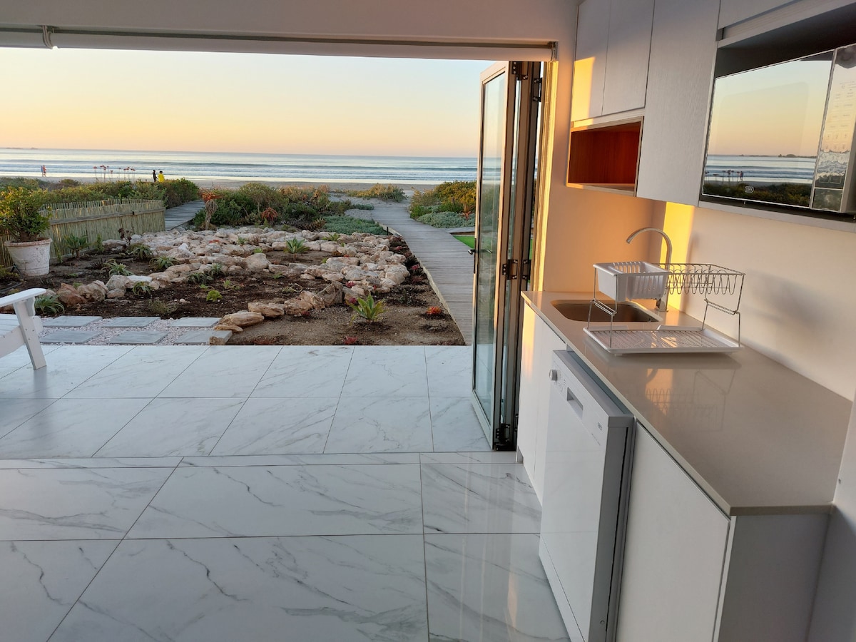 A modern kitchen with sleek cabinetry and a clean countertop is visible. Glass doors open to reveal a serene beach view, while a well-maintained pathway leads to the water. Soft morning light enhances the tranquility of the space.