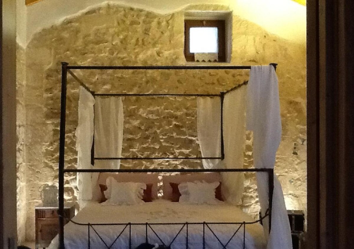 A canopied bed is framed by soft white drapes in a cozy sleeping area. The stone walls contribute a rustic feel, while natural light enters through a small window above. Two decorative pillows enhance the inviting appearance of the spacious bed.