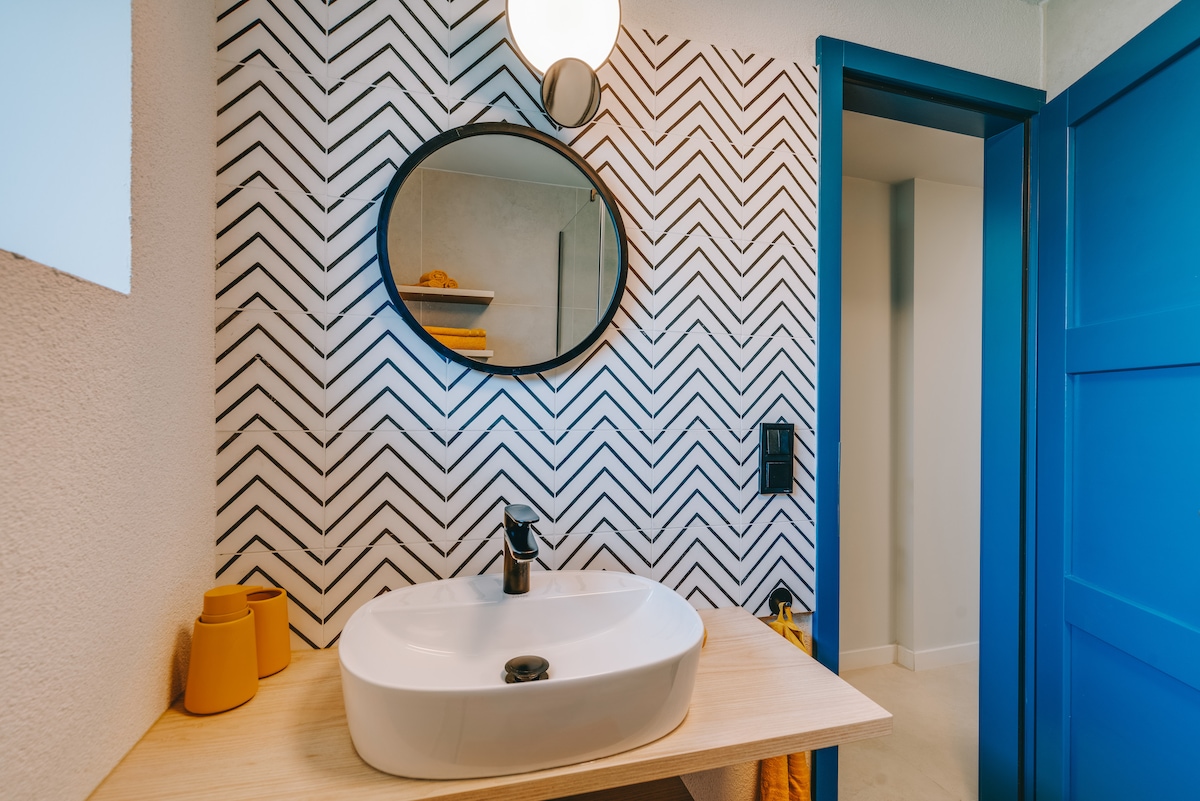 A modern bathroom features a circular mirror above a white sink on a wooden countertop. Bold chevron-patterned wallpaper creates a striking backdrop. Brightly colored accessories, including yellow cups and towels, add a splash of color. A blue door provides a contrasting element to the space.