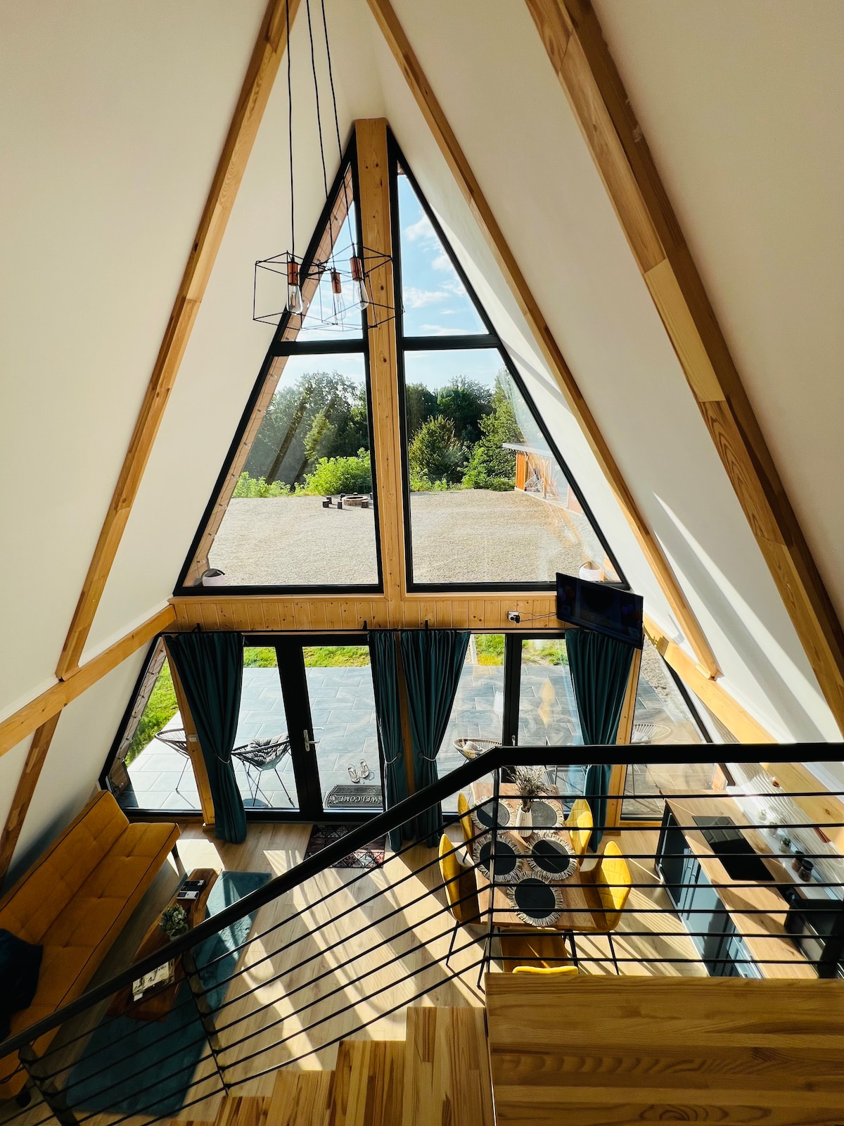 An aerial view captures the open concept living area with large triangular windows. Natural light floods the space, showcasing wooden elements and modern furnishings. A staircase leads to an inviting seating area below, with a lounge chair positioned beside a large glass door opening to outdoor greenery.