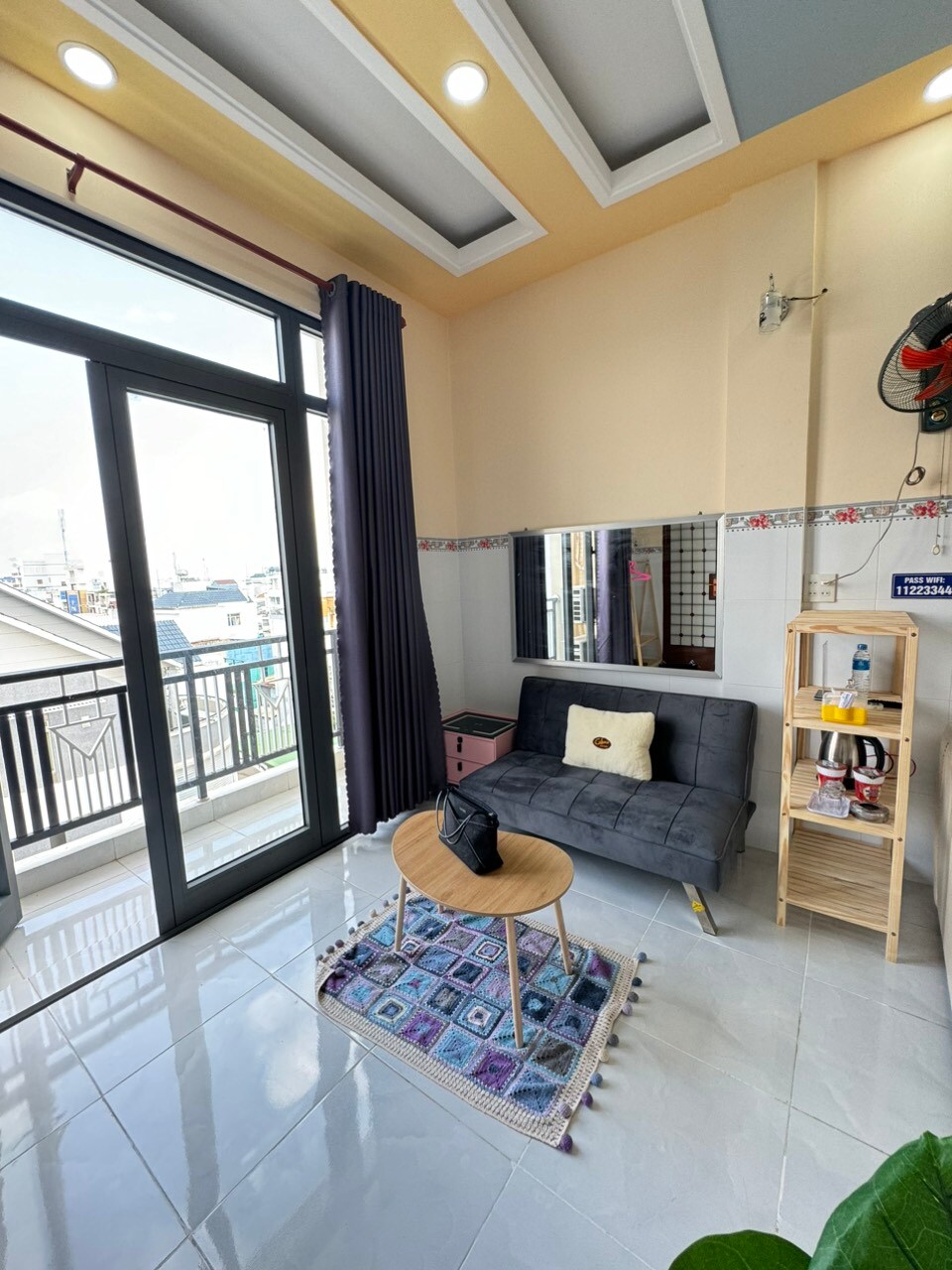 A bright living space is shown with large glass doors allowing natural light to enter. A comfortable sofa bed faces a small round table on a decorative rug, while a shelving unit holds various items. Neutral tones enhance the room's airy atmosphere.