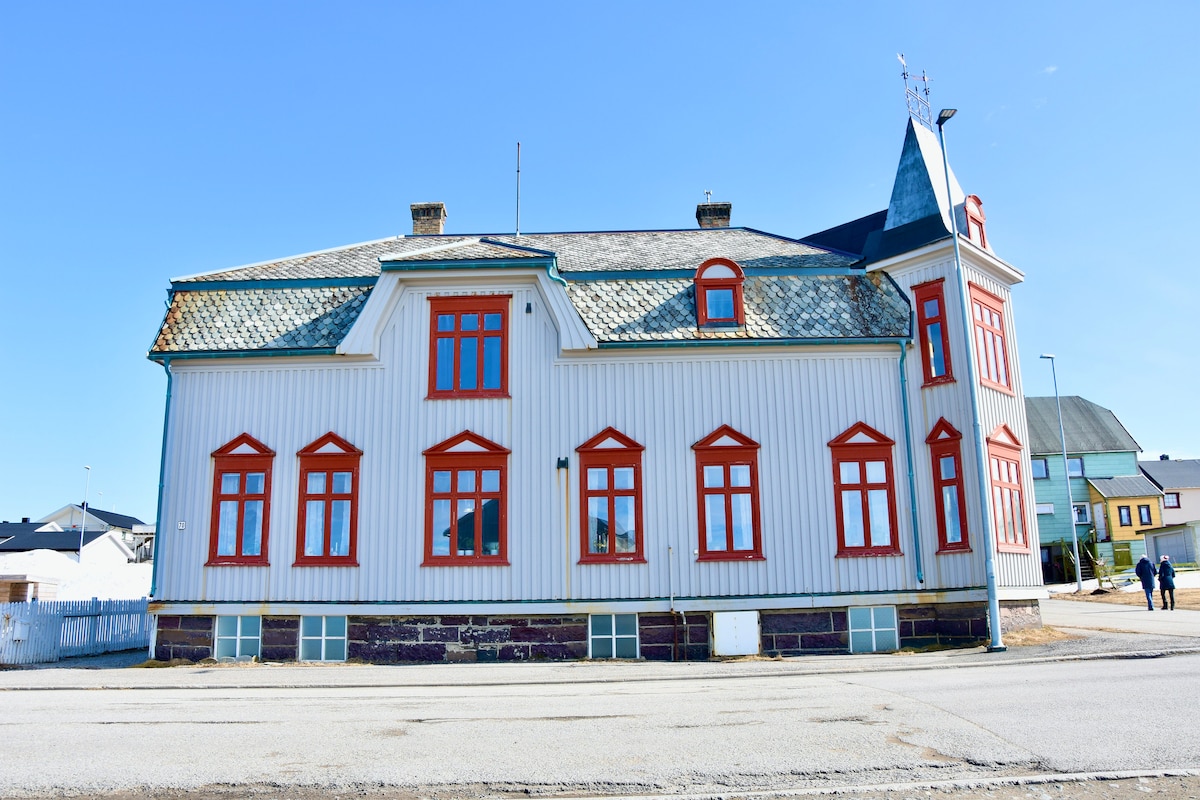 The historic building features a mix of traditional architecture with a striking white exterior and bright red window frames. A distinctive turret and steeply pitched roof are visible, contributing to the building's unique character. The surrounding area shows paved access and neighboring structures.