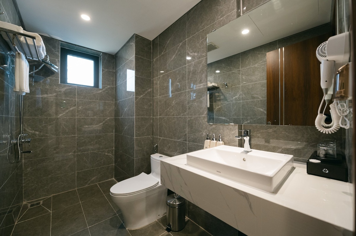 The bathroom features contemporary grey marble walls and flooring. A sleek white sink is positioned on a modern countertop, complemented by a mirror above. A toilet is situated beside the sink, with a window allowing natural light. Towel racks and a hairdryer offer added convenience.