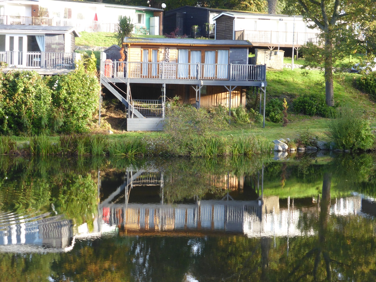 A detached chalet is positioned near a tranquil lake, featuring spacious decking and large windows. The wooden structure reflects on the water, surrounded by greenery. An outdoor area is visible, creating a serene setting for relaxation.