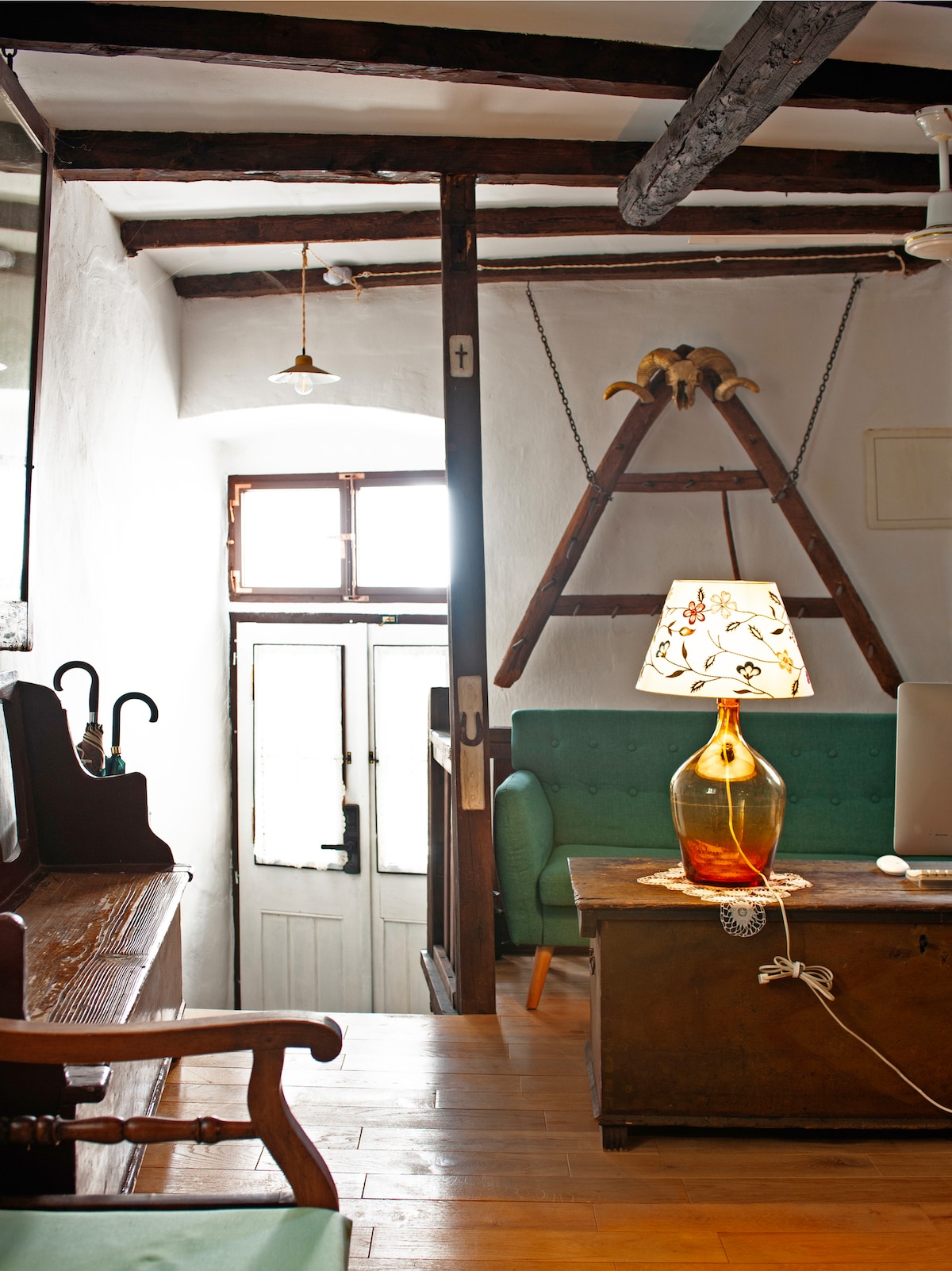 A cozy living space is shown, featuring wooden beams and warm natural light. A green sofa is positioned near a wooden table, which holds a decorative lamp. Traditional artifacts, such as a wooden ladder and vintage furniture, enhance the historical charm of the room.