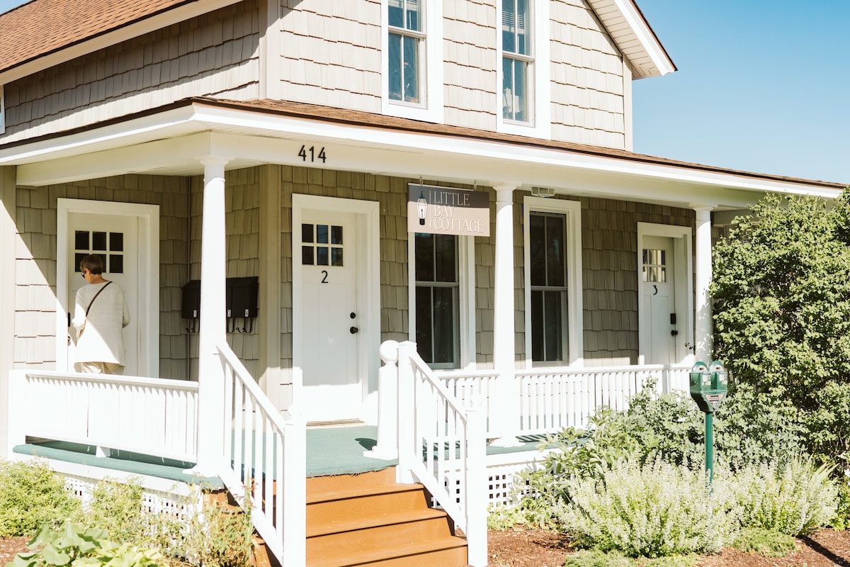 The exterior of Little Bay Cottage is showcased, featuring a two-story structure with a light gray shingle facade. The welcoming entrance is complemented by white railings and stairs. Numbered doors are visible, indicating individual units, with lush greenery surrounding the cottage.