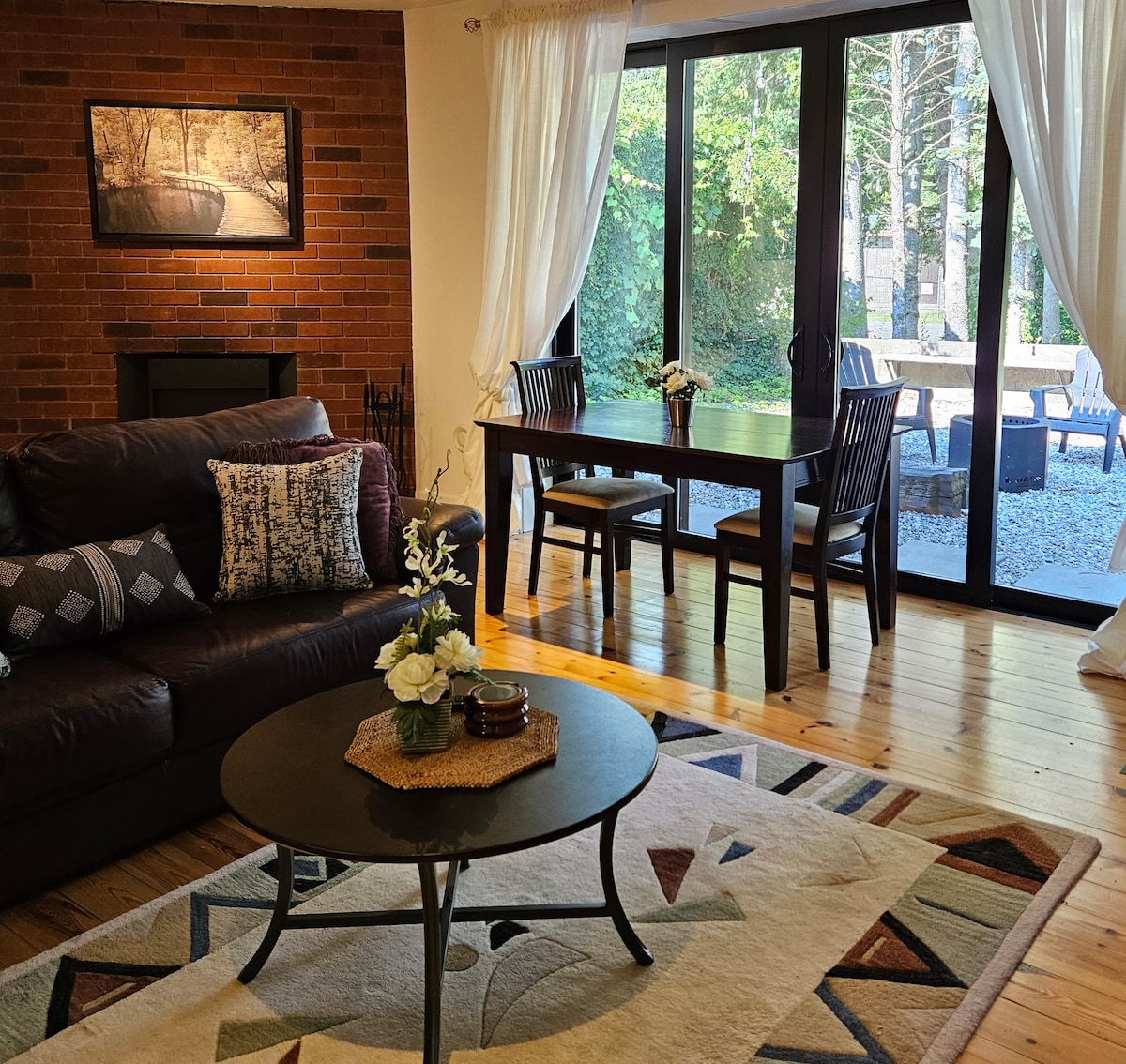 A comfortable living area features a dark leather sofa adorned with patterned cushions. A round coffee table is set with decorative items. The space is brightened by large sliding glass doors leading to an outdoor seating area, surrounded by greenery.