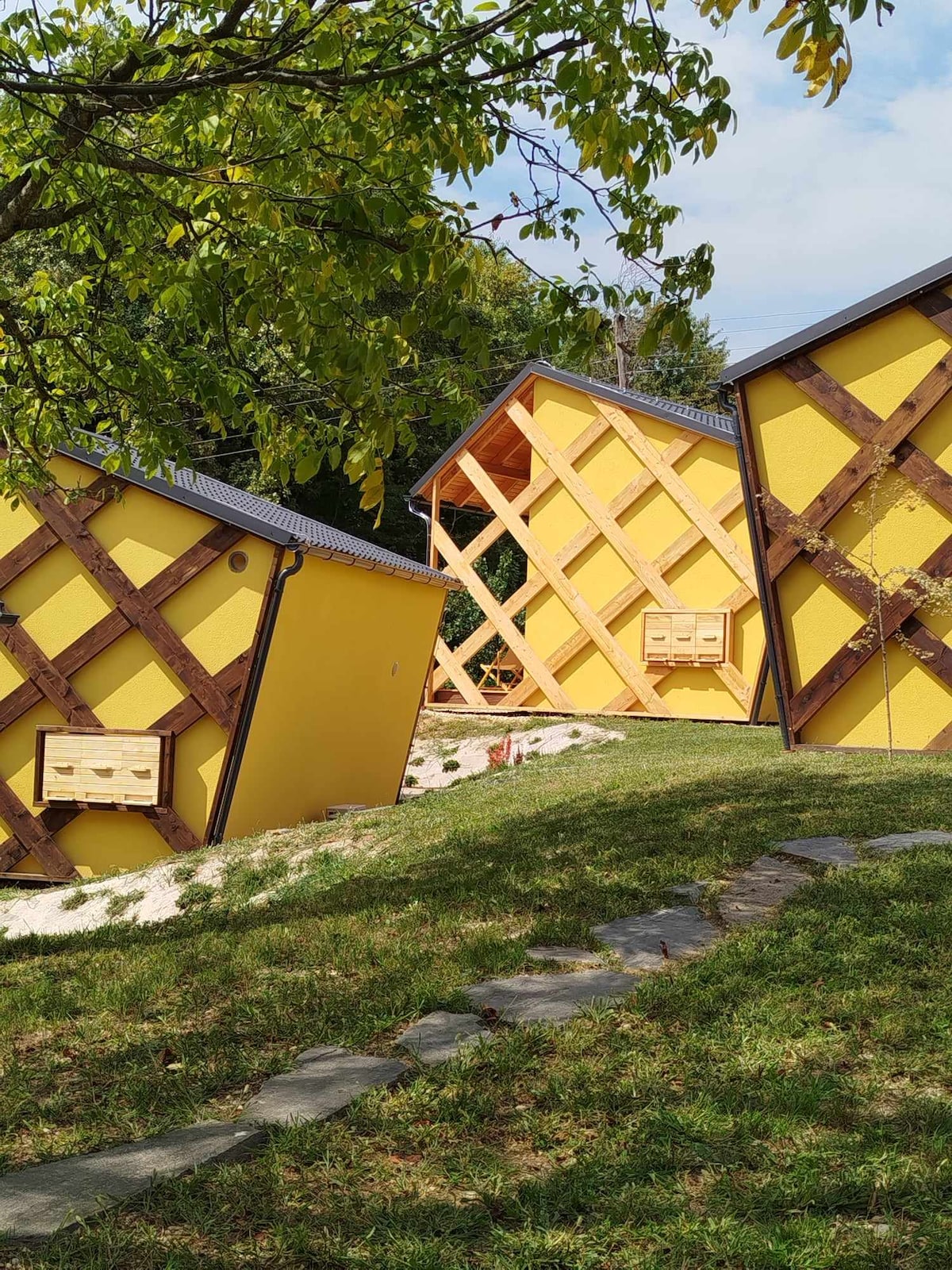Two yellow cottages are viewed from the side, set against a gentle hillside. Each cottage features wooden accents and bee hives integrated into the design. A stone path leads through the lush green grass, inviting guests to explore the surrounding area.