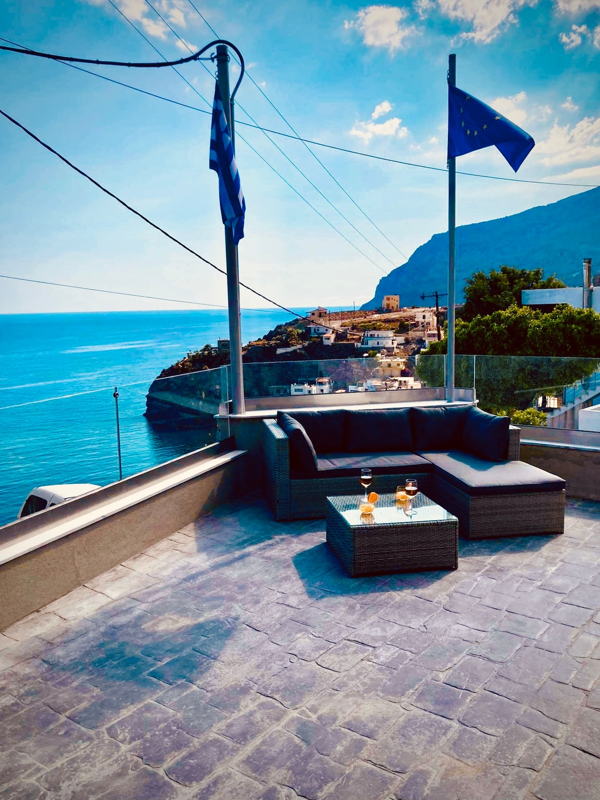 A spacious outdoor terrace is presented, furnished with a modern seating arrangement overlooking a breathtaking coastal view. Two glasses filled with a beverage rest on the glass-topped table, with flags gently fluttering in the breeze against a backdrop of blue water, cliffs, and sky.