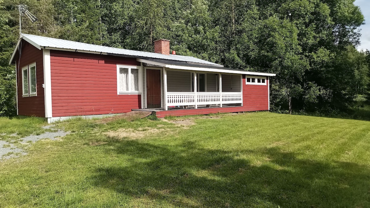 A red wooden house is set amidst lush greenery, featuring a spacious front porch with white railings. The building has multiple windows, allowing natural light to illuminate the interior. A well-kept lawn surrounds the house, providing a serene outdoor space.