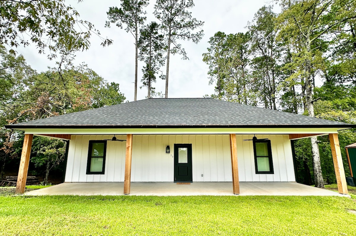 The front exterior of the cabin features a covered porch with wooden pillars, complementing the white siding and dark roof. The entrance is marked by a green door, flanked by black outdoor light fixtures. Lush green grass surrounds the porch, enhancing the natural setting.