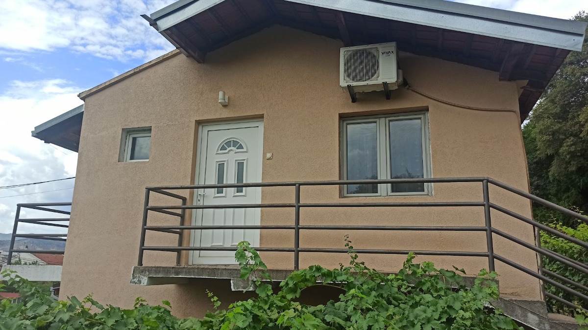 A light-colored building features a welcoming entrance with a white door and a stylish railing. Air conditioning is visible on the wall, and large windows allow natural light to fill the space. Green foliage surrounds the base, offering a touch of nature.