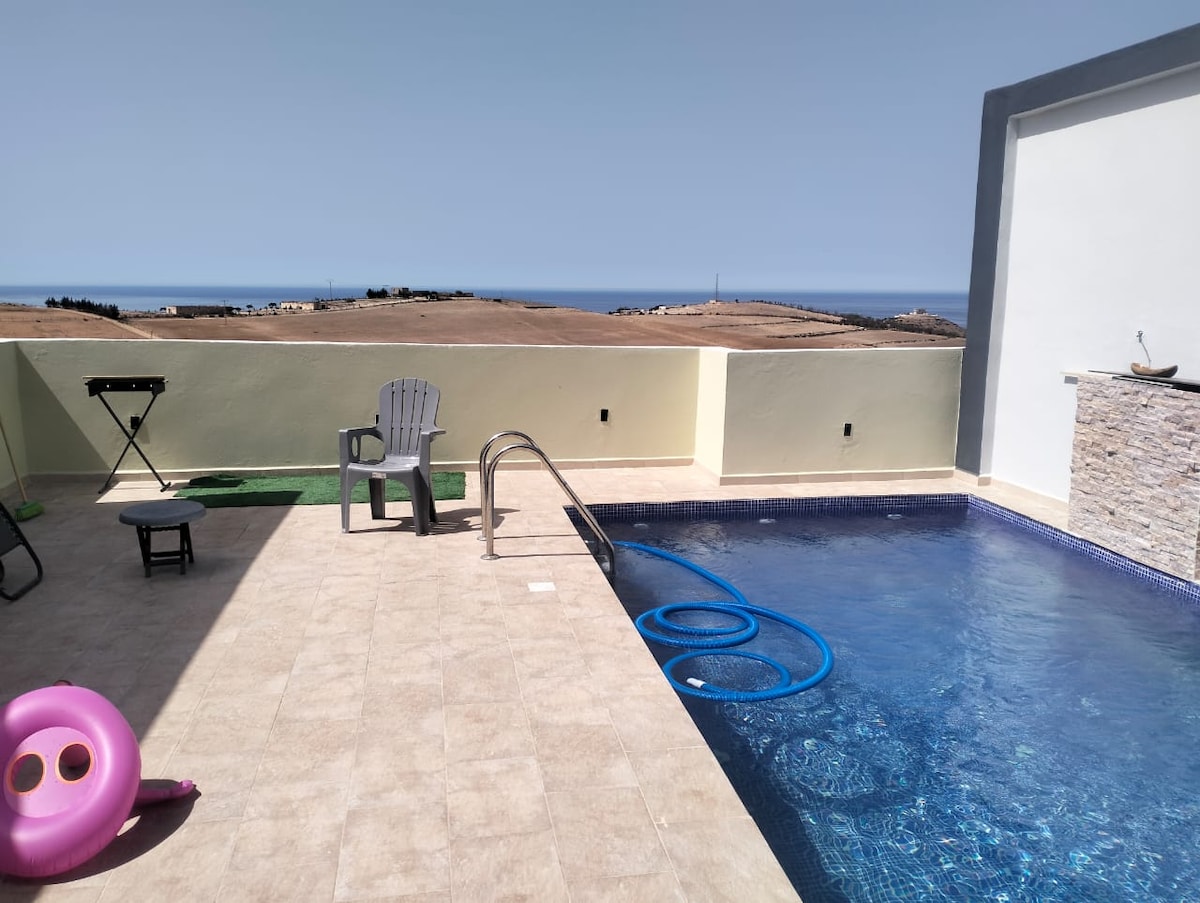 An outdoor pool area is presented, featuring a clear blue pool with a ladder for easy access. A small table and a couple of chairs are positioned nearby, along with a BBQ grill. Scenic hills and a distant view of the coastline can be observed in the background.