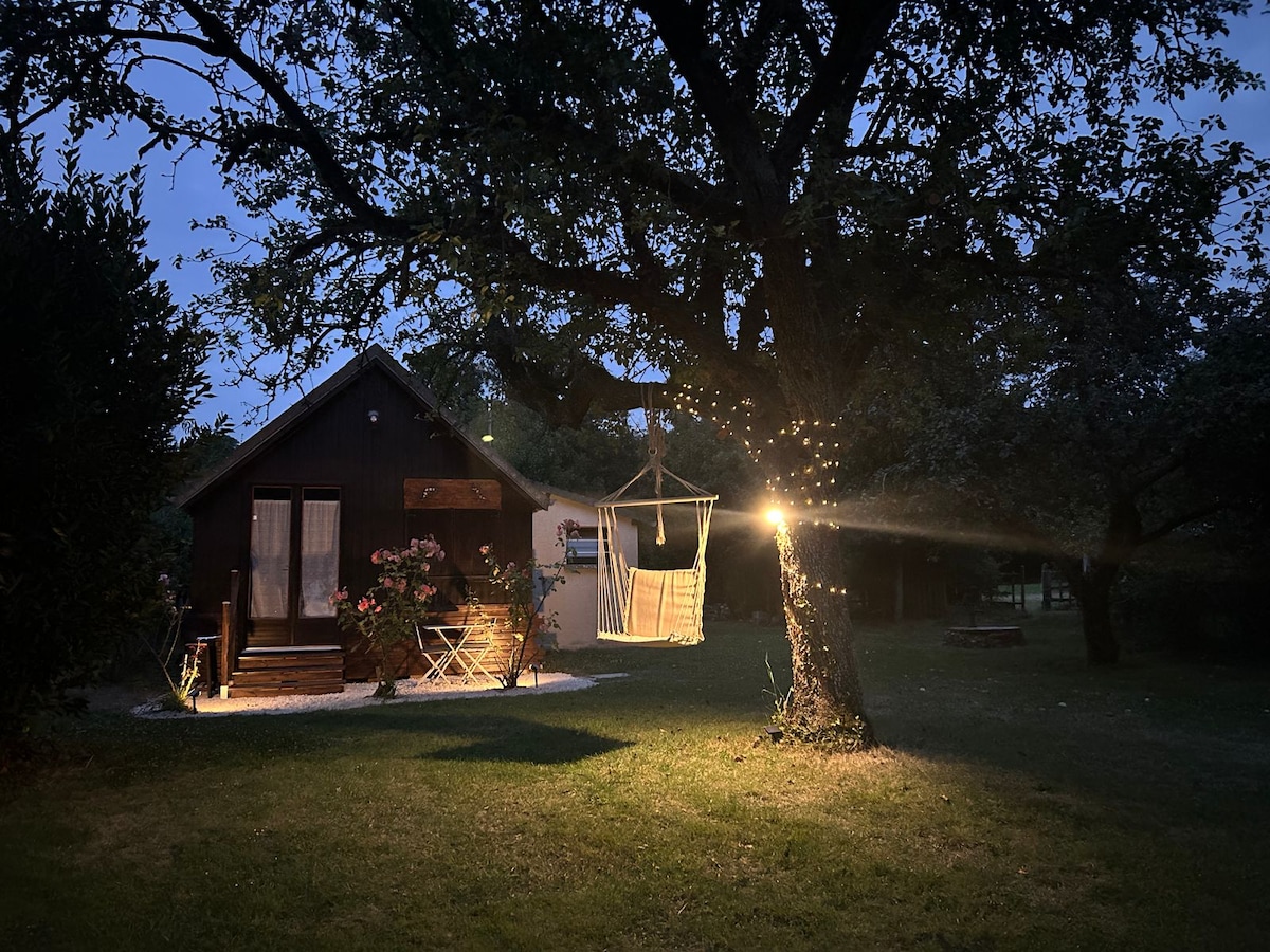 A charming chalet is nestled among trees, with warm lighting illuminating the exterior. A hammock hangs from a sturdy tree, offering a relaxed seating option. The surrounding grassy area is dotted with trees and garden features, creating a tranquil outdoor setting.