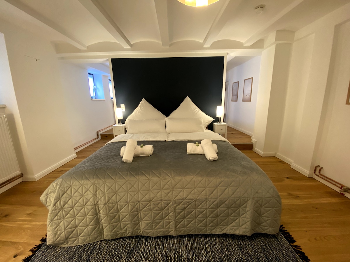 The sleeping area features a spacious bed with matching decorative pillows and a soft gray quilt. Nightstands with lamps are placed on either side. Light is filtered through windows, illuminating the warm wood flooring and minimalistic decor.