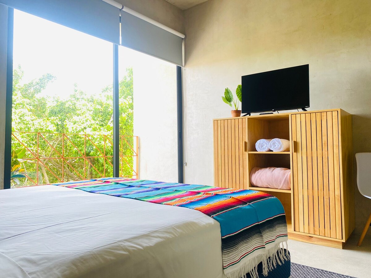 A spacious bedroom is furnished with a large bed covered by a vibrant striped blanket. A wooden cabinet holds neatly stacked towels and a TV is positioned on top. Large windows offer views of greenery, allowing natural light to fill the room.