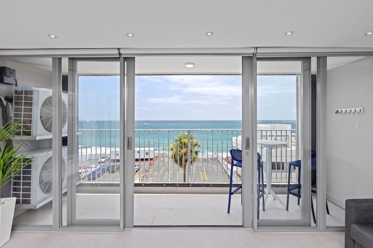 The image presents a spacious balcony with sliding glass doors that open to an ocean view. A small table and two chairs are positioned for outdoor seating. The scene captures the vibrant blue ocean and a glimpse of the parking area below.