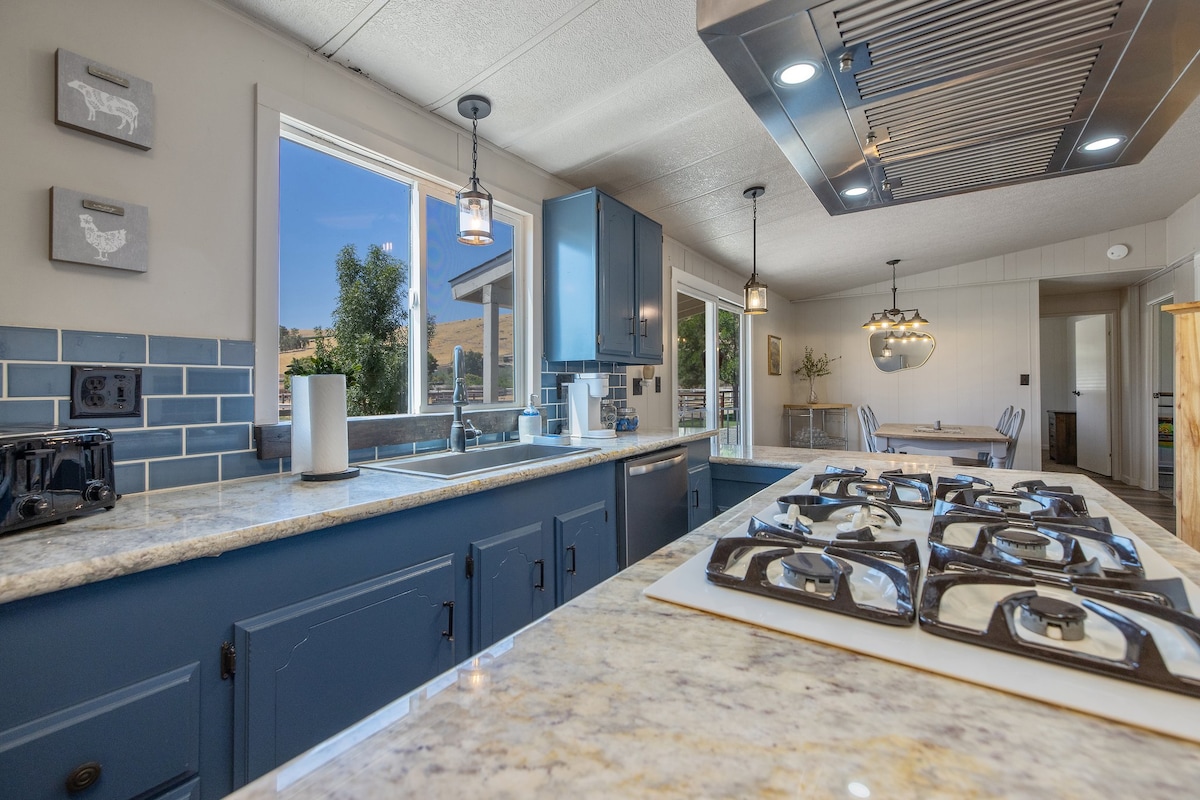 A modern kitchen space is presented, featuring granite countertops and a gas stove with multiple burners. Blue cabinetry complements the light blue tile backsplash. Large windows allow natural light to fill the area, offering views of the outdoors, with a dining area visible in the background.
