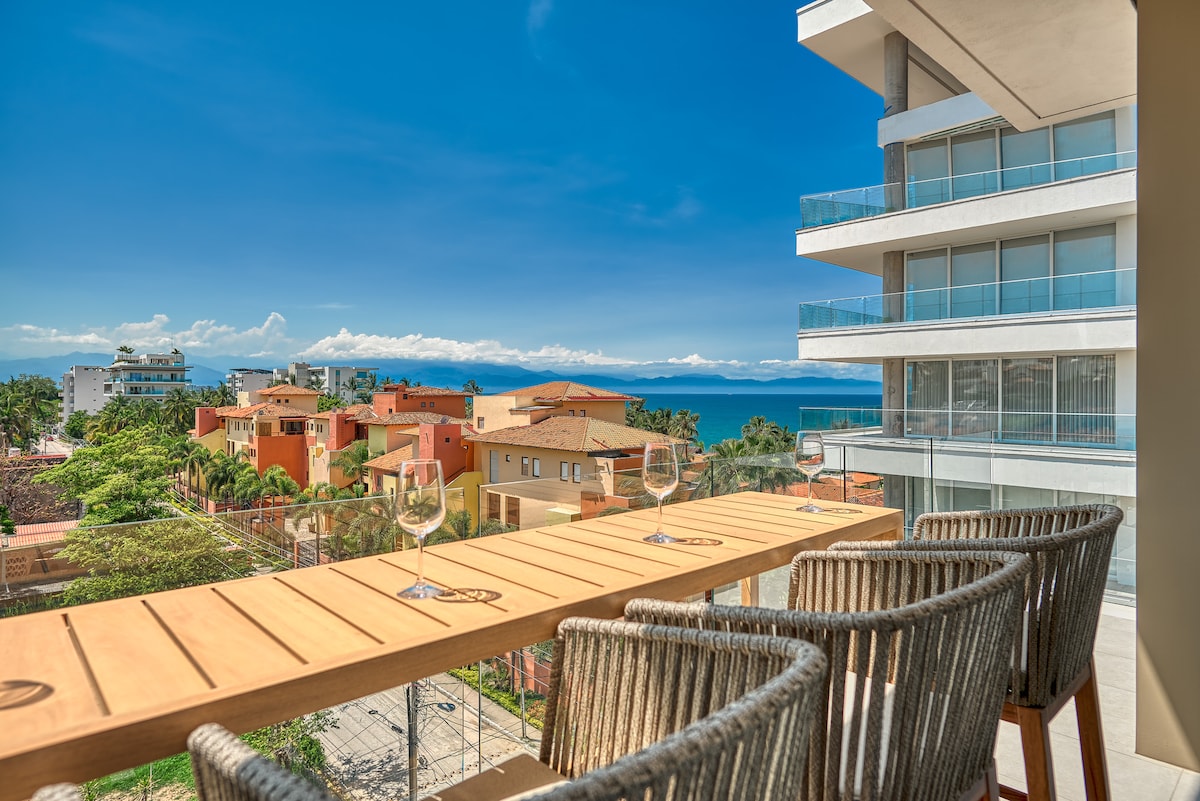 An outdoor balcony features a wooden bar accented by four woven chairs, overlooking a vibrant town and the blue ocean. The clear sky and distant mountains complete the scenic view, inviting relaxation and enjoyment of the surroundings.