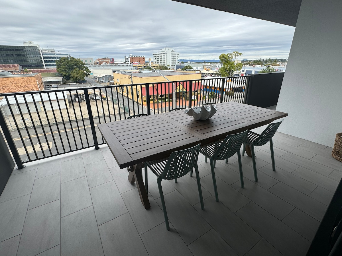 A spacious outdoor balcony features a large wooden table accompanied by several green chairs. The area is equipped for gatherings and offers views of the surrounding buildings and distant water. A neutral color palette enhances the modern aesthetic of the space.