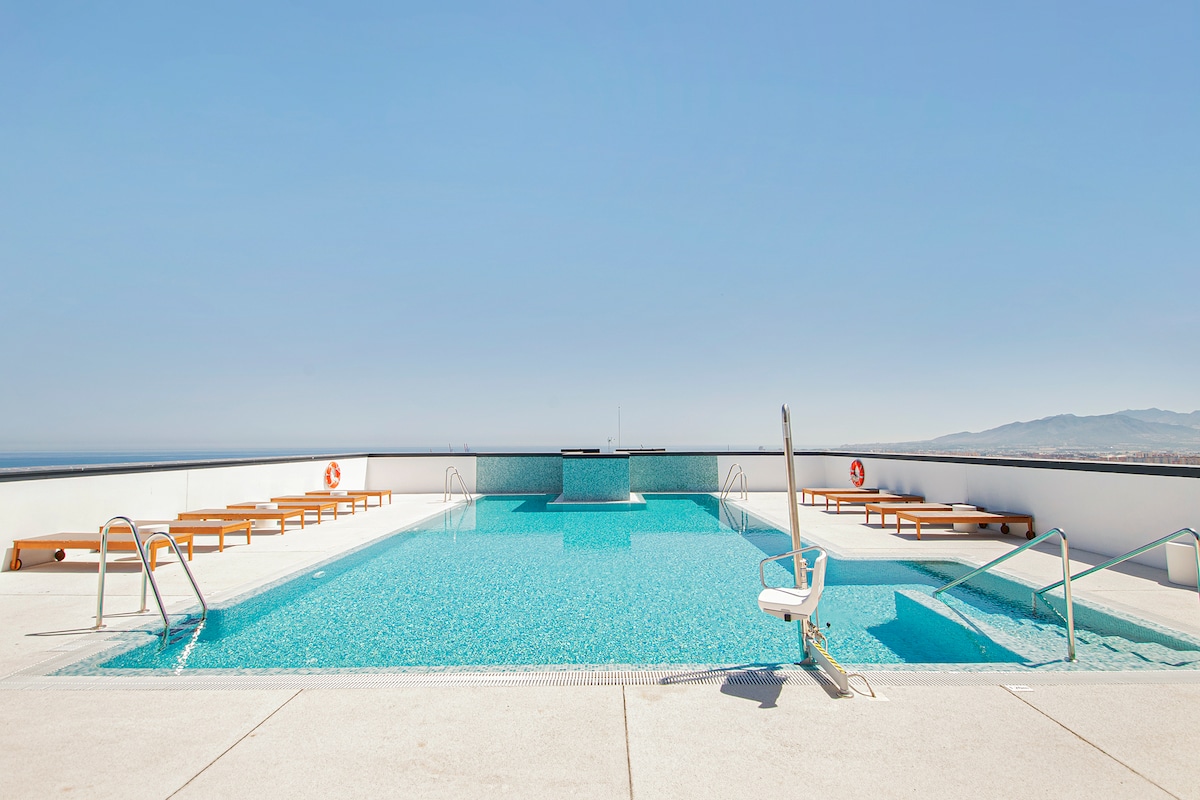 A rooftop pool with a clear water surface occupies the upper level, offering sweeping views of the city and horizon. Surrounding the pool, wooden loungers provide relaxation options. Safety equipment, including a life ring, is positioned strategically for guest security.