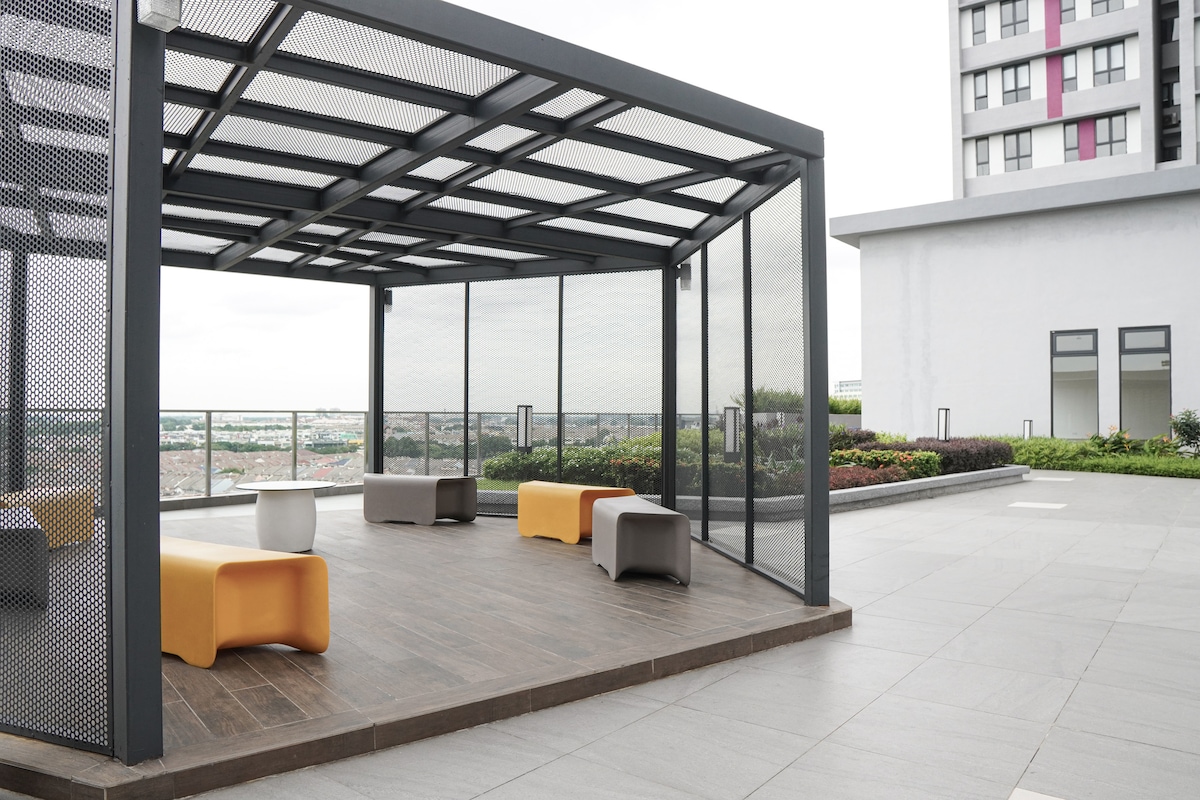 A covered lounge area features modern, modular seating options in neutral tones, complemented by an accent piece in orange. The wooden deck offers a warm contrast to the contemporary metal structure, with a landscaped area visible beyond, providing a serene environment.