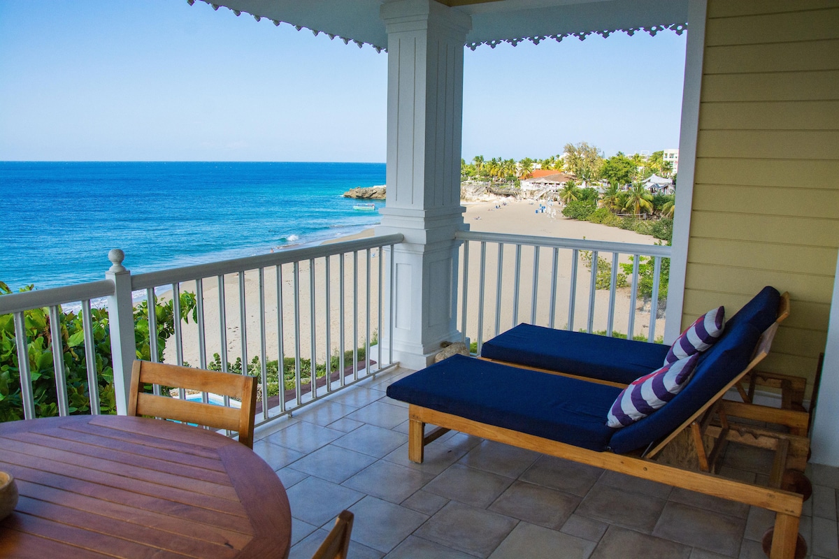 A spacious balcony is furnished with a round wooden table and two lounging chairs, featuring striped cushions. The oceanfront view showcases calm blue waters meeting the sandy beach, framed by lush greenery and distant shoreline, inviting relaxation and enjoyment of the scenic landscape.
